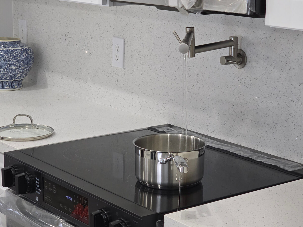 Pot filler faucet running water into pot on induction cooktop with quartz backsplash