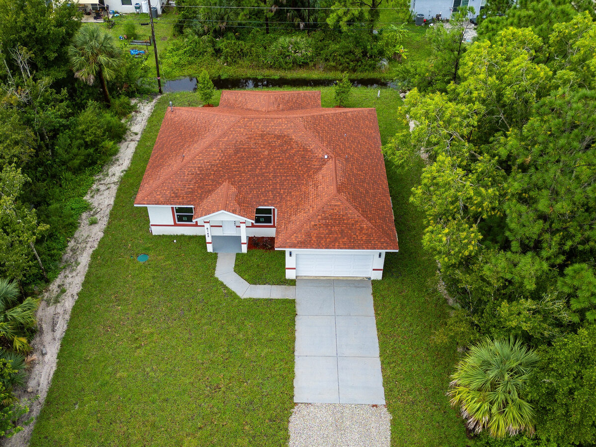 Drone aerial of full property — shingled roof, green lawn, concrete driveway and mature trees